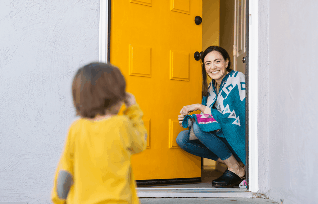 A woman kneels and smiles at a small child standing outside a bright yellow door in Uplands Westminster. The woman holds a blanket as they share a warm moment at the entrance of their home.