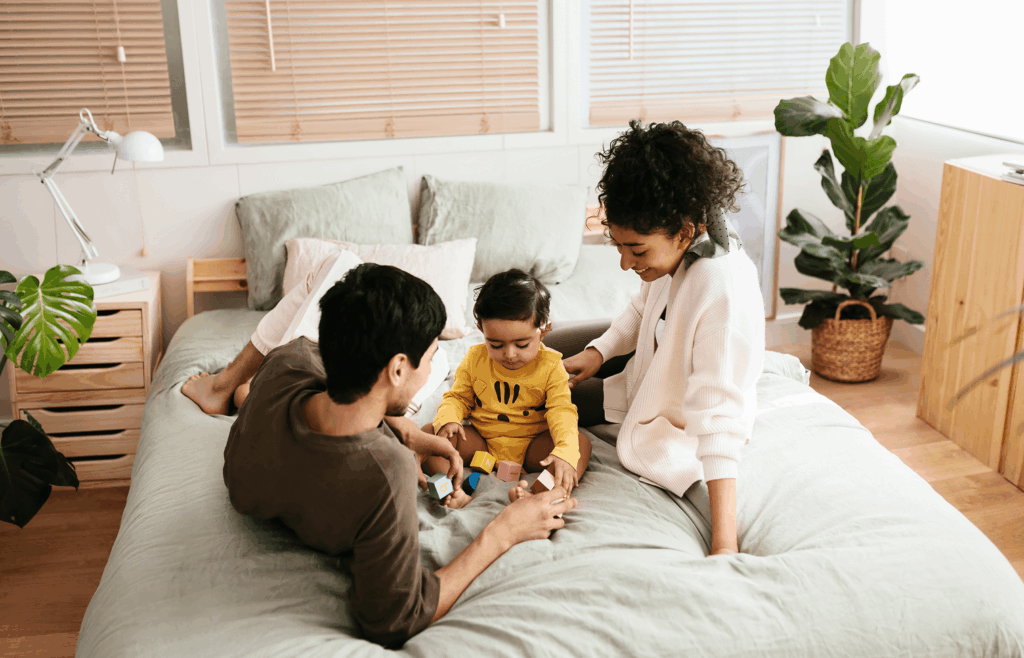 Two adults and a toddler sit on a bed in a bright, cozy bedroom, playing with wooden blocks—a charming scene often found in Uplands Westminster homes for sale.