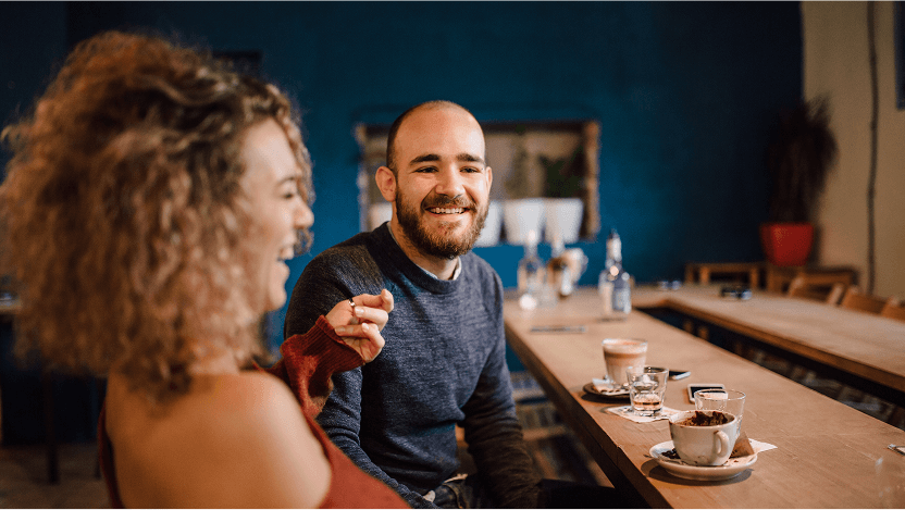 Two people sit at a wooden table in a café in Uplands, sharing drinks and dessert. One person laughs while the other smiles, enjoying coffee as they chat about homes for sale in Westminster CO.