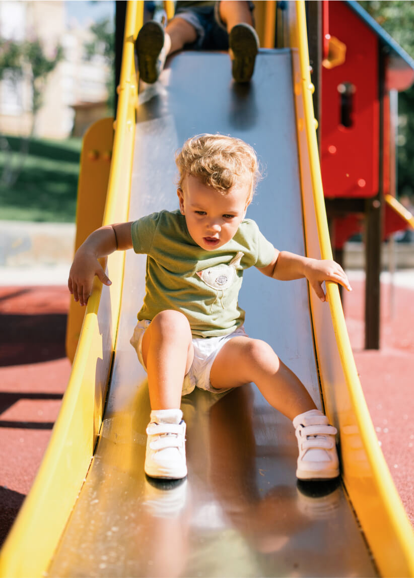A young child in a green shirt and white shoes sits on a yellow playground slide in Uplands Westminster, preparing to go down, with another child’s legs visible at the top.
