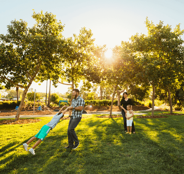 A man swings a child by the arms while a woman and another child stand nearby in a sunlit park with trees and grass, capturing joyful moments near Uplands Westminster homes for sale.