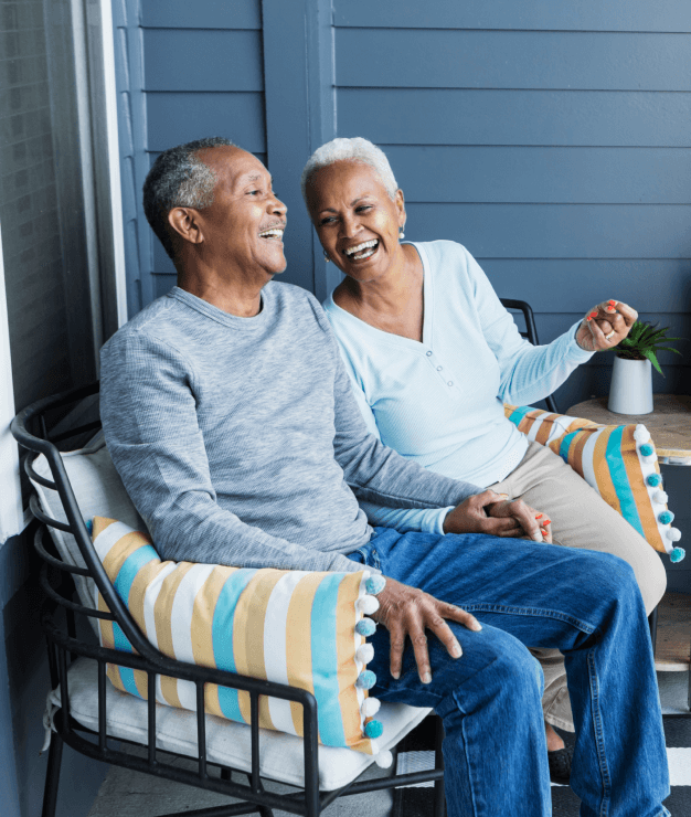 An older couple sits together on a porch at Uplands Westminster, smiling and holding hands, with striped pillows on their chairs and a small table nearby. Discover the warmth of homes for sale in Westminster CO.