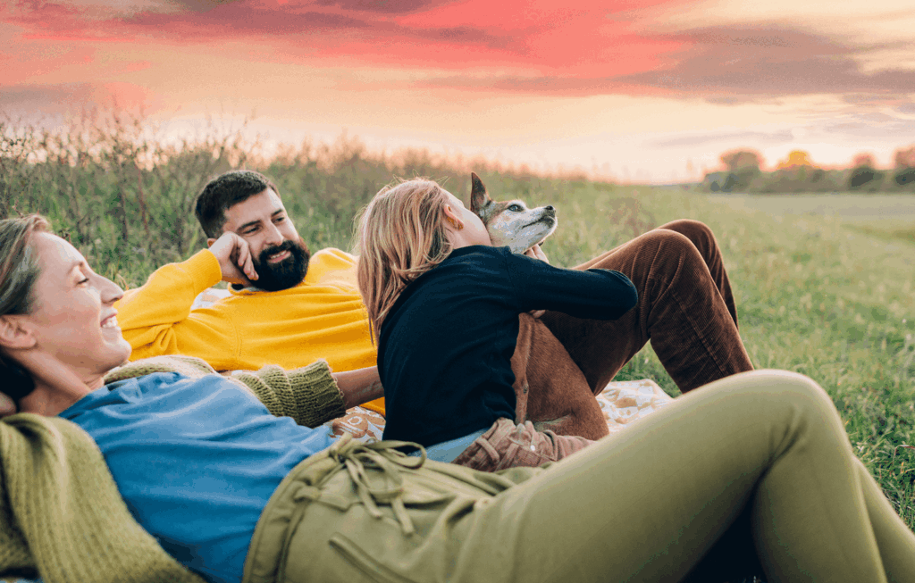 Three people relax on a blanket in a grassy field at sunset while one person hugs a dog, enjoying the serene beauty near Uplands Westminster homes for sale.