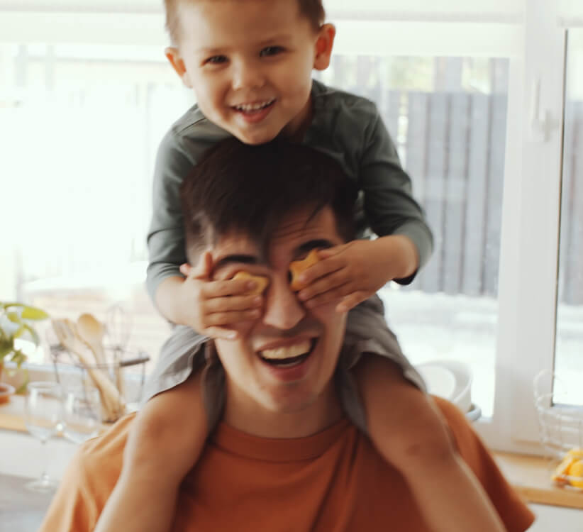 A smiling child sits on an adult's shoulders, playfully covering the adult's eyes with their hands in a bright kitchen—capturing a joyful moment in one of the beautiful Uplands Westminster homes for sale.