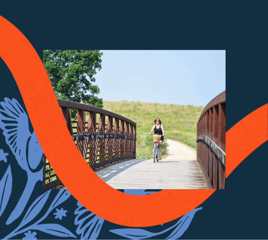 A person rides a bicycle across a wooden bridge in a grassy, open area beneath a clear sky, with graphic orange and blue design elements. Explore the charm of Uplands Westminster and discover homes for sale in Westminster CO.