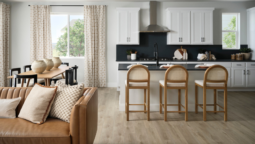 Modern kitchen and dining area in Uplands Westminster features white cabinets, black backsplash, an island with three barstools, dining table with vases, and a tan sofa with patterned pillows—perfect for those seeking homes for sale in Westminster CO.