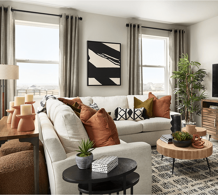 A modern living room at Uplands Westminster features a white sectional sofa, throw pillows, potted plants, a round wooden coffee table, abstract wall art, and two large windows with gray curtains—perfect for enjoying Uplands Westminster homes for sale.
