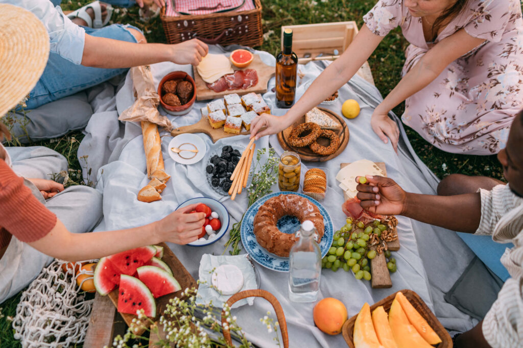 Several people sit on a blanket outdoors, enjoying a picnic with bread, fruit, cheese, and pastries. Plates, a wine bottle, and a basket are in view—capturing the relaxed lifestyle near Uplands Westminster homes for sale.
