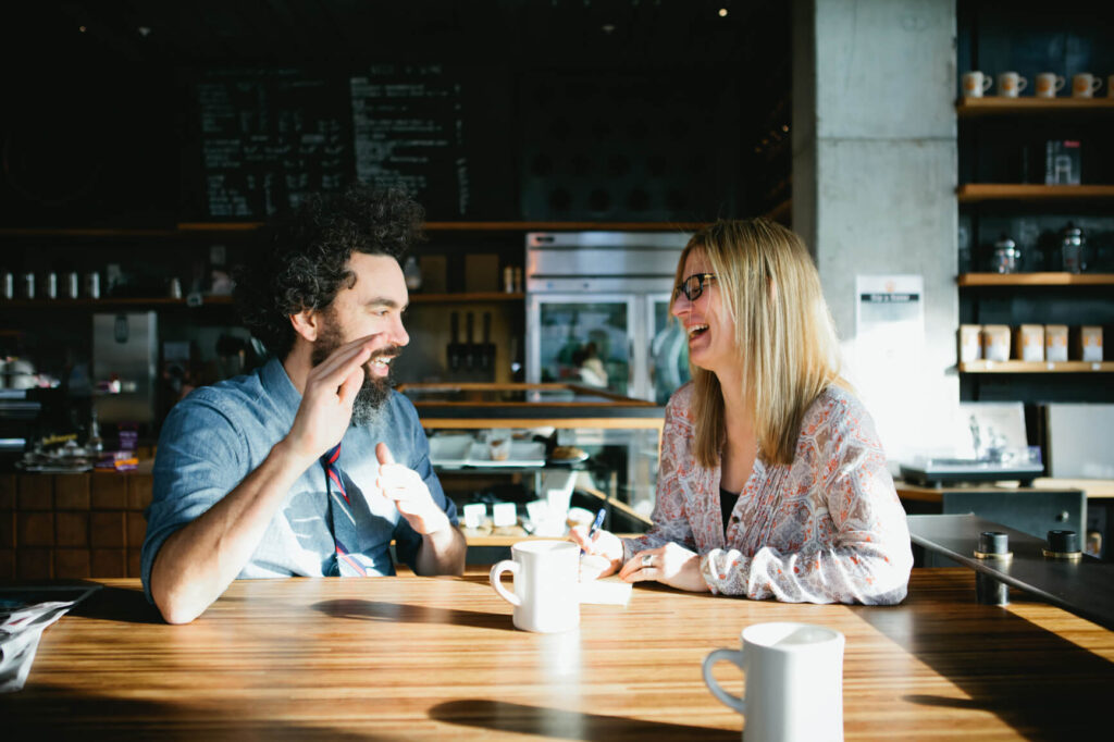 Two people sit at a wooden table in a sunlit café, smiling over coffee mugs, chatting about Uplands Westminster and the best homes for sale in Westminster, CO.