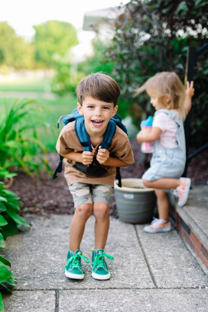 A young boy with a backpack smiles at the camera while a younger child sits nearby holding a cup in an outdoor garden, capturing the welcoming spirit found near Uplands Westminster homes for sale.