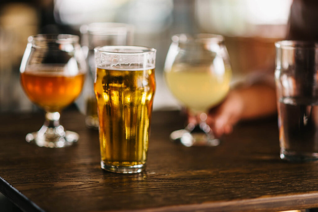 A pint glass of beer sits on a wooden table next to several goblets with different drinks and a glass of water, with a blurred hand in the background—a cozy Uplands Westminster vibe perfect for relaxing after exploring Uplands Westminster homes for sale.