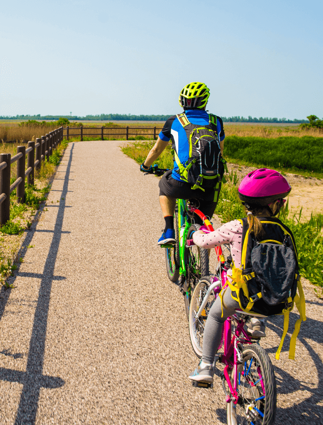 An adult and a child, both wearing helmets and backpacks, ride bicycles on a paved path through the scenic Uplands Westminster countryside, enjoying the peaceful surroundings near beautiful Uplands Westminster homes for sale.