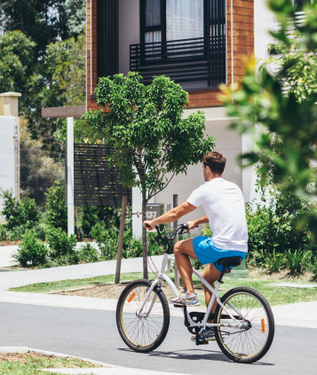 A person wearing a white shirt and blue shorts rides a bicycle on a paved path in Uplands, passing by modern houses and greenery, showcasing the inviting atmosphere near Uplands Westminster homes for sale.