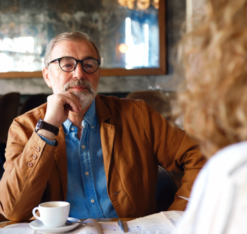 An older man with glasses and a brown jacket sits at a table with a cup of coffee, looking thoughtfully at a person across from him, perhaps discussing life in Uplands or sharing thoughts on Uplands Westminster homes for sale.