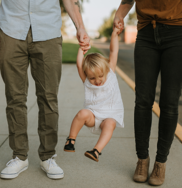 A child in a white dress is being swung by the arms by two adults, one on each side, while walking on a sidewalk in the scenic Uplands neighborhood, known for its beautiful Westminster homes for sale.