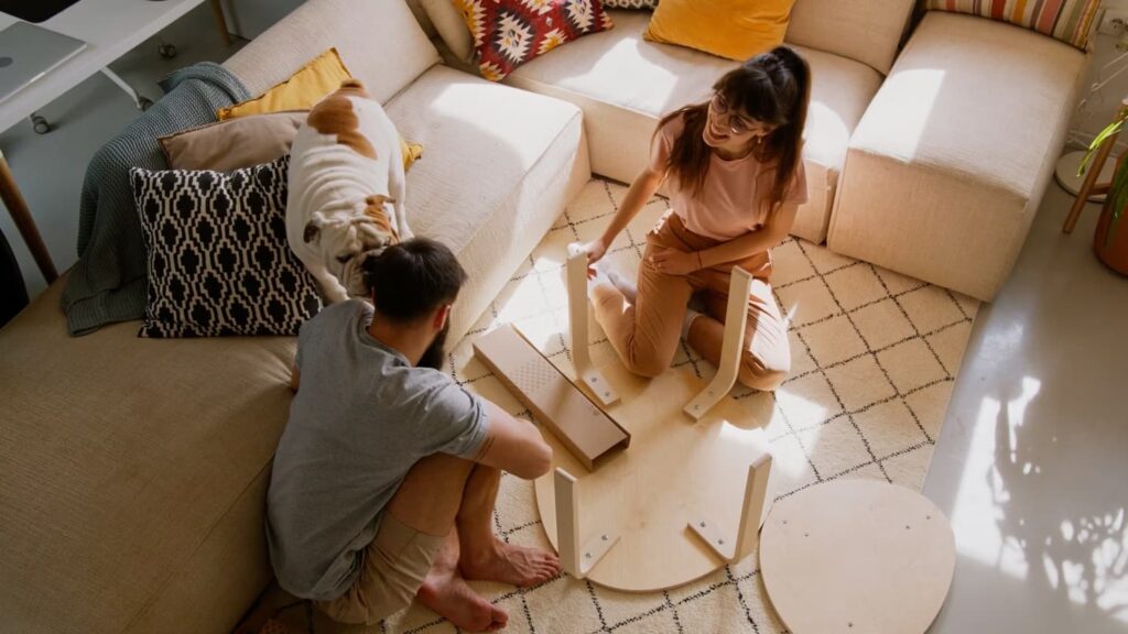 Two people assemble a round wooden table on a patterned rug in a living room, while a dog sits on the couch nearby.