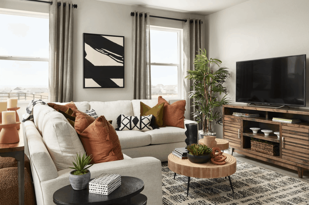 A modern living room in Uplands Westminster features a white sectional sofa, orange pillows, a round wooden coffee table, potted plants, a TV on a wooden stand, and two windows with gray curtains—perfect for those seeking homes for sale in Westminster CO.