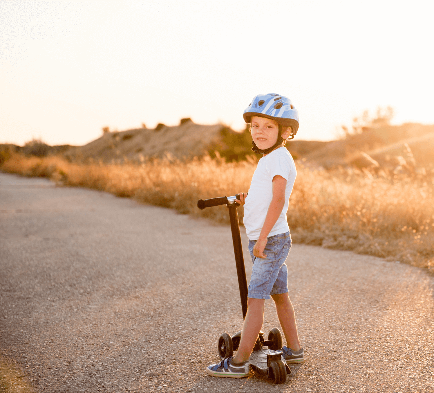 Young child wearing a blue helmet stands on a scooter along a paved path at sunset, with dry grass and hills in the background—capturing the outdoor spirit near Uplands Westminster homes for sale.