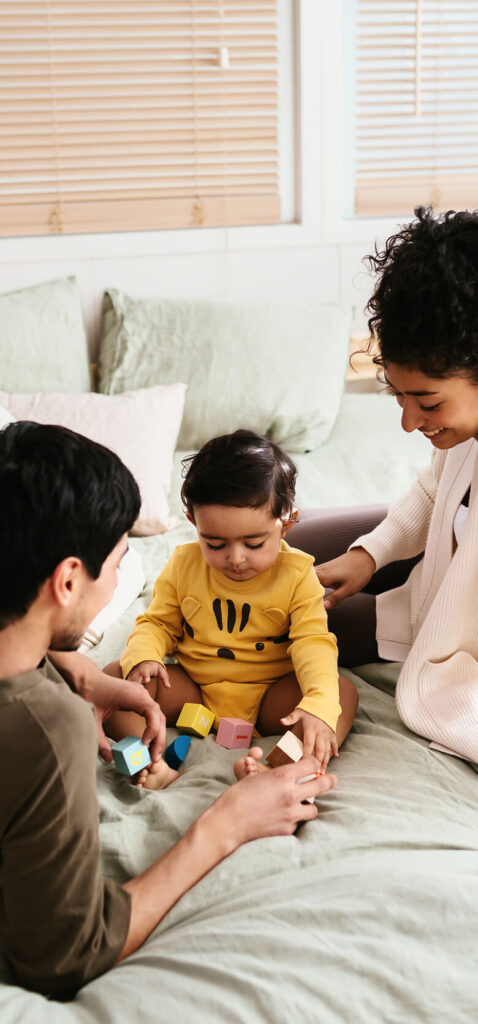 Two adults sit on a bed with a toddler dressed in yellow, playing with colorful wooden blocks—a cozy family moment that captures the warmth of living in Uplands Westminster, where inviting homes for sale in Westminster CO await.
