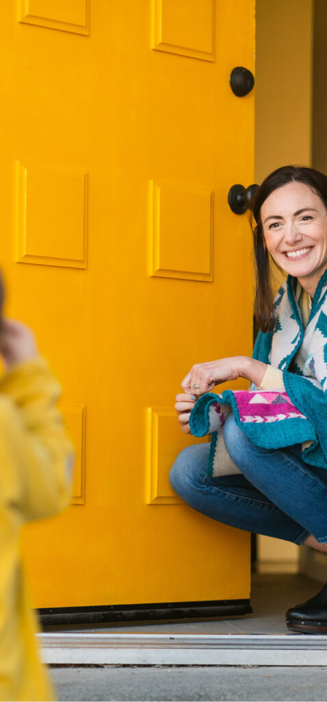 A woman kneels and smiles at a child standing outside as she holds open a bright yellow door, welcoming them into an inviting Uplands Westminster home for sale.