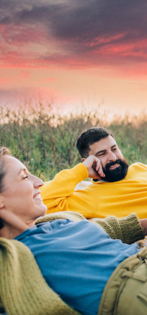 Two people lying on the grass outdoors at sunset, smiling and relaxing amid lush greenery and a colorful sky—capturing the tranquil charm of Uplands Westminster.